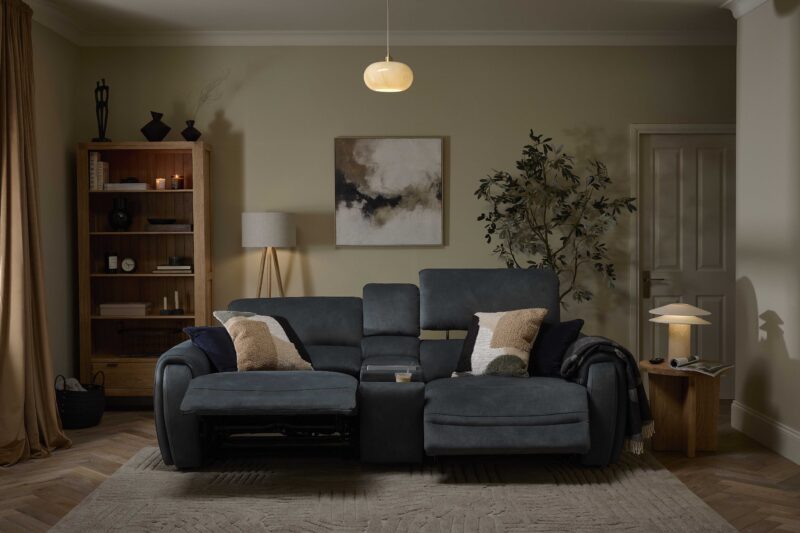 An sofa and side table-living room furniture-reclining dark blue sofa-round wooden side table-neutral textured rug-parquet floor-tray with books-modern cream table lamp-neutral walls-off white door-large house plant-abstract neutral painting-floor standing lamp-wooden bookcase-draped curtains-sculptural black vases- glass ceiling light