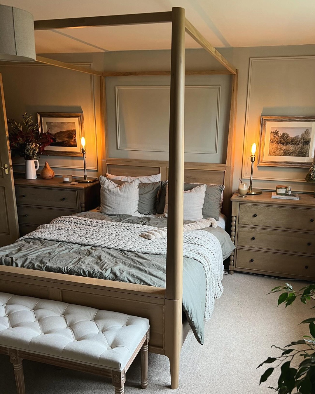 A bed and chest of drawers-wooden four poster bed-cushioned bench-neutral bed linen-wooden chest of drawers-wall beading-sage green walls-candle light-framed landscapes-beige carpet