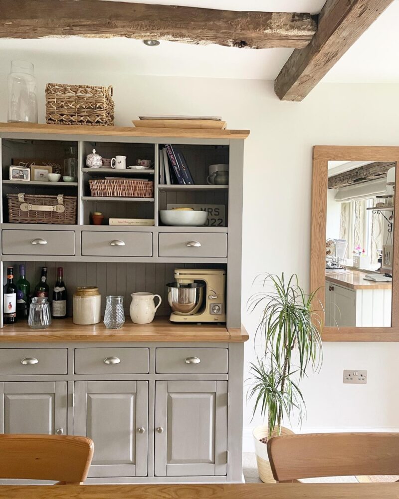 A dresser, mirror, and dining table-kitchen dining furniture-a grey painted large dresser-wooden rectangular mirror-wooden table and chairs-food mixer-storage jars and baskets-house plant-exposed beams-off white walls