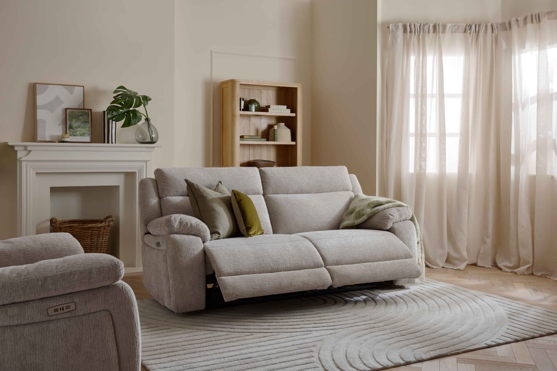 A sofa, bookcase, and armchair-neutral reclining sofa-sheer draped curtains-wooden bookcase-neutral textured rug-parquet wooden floor-neutral reclining armchair-off white fireplace-off white walls-woven storage basket