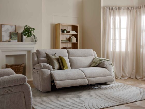 A sofa, bookcase, and armchair-neutral reclining sofa-sheer draped curtains-wooden bookcase-neutral textured rug-parquet wooden floor-neutral reclining armchair-off white fireplace-off white walls-woven storage basket