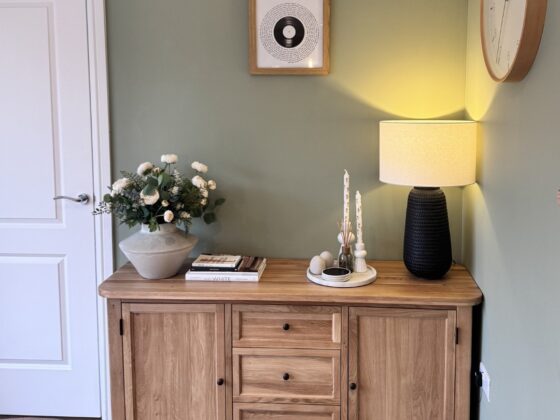 A sideboard and door-dining room furniture-large wooden sideboard-black table lamp-grey vase with fresh flowers-tray with tapered candles-stacked books-wooden wall mounted clock-framed wall art-green walls-white door-wooden floor-wooden dining table-grey upholstered dining chair