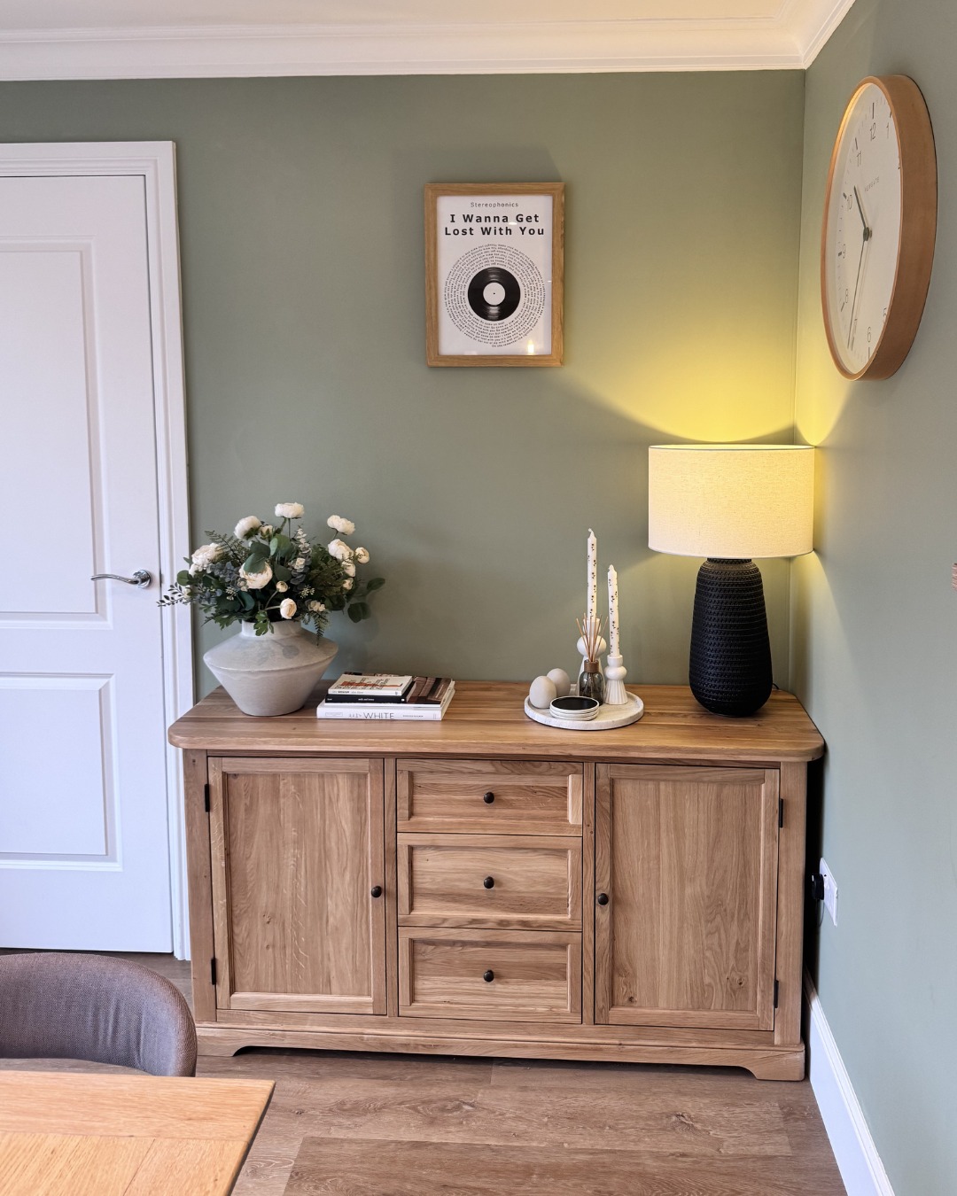 A sideboard and door-dining room furniture-large wooden sideboard-black table lamp-grey vase with fresh flowers-tray with tapered candles-stacked books-wooden wall mounted clock-framed wall art-green walls-white door-wooden floor-wooden dining table-grey upholstered dining chair