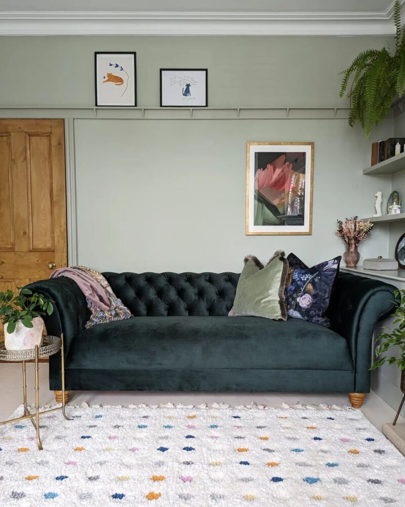 A sofa-living room furniture-plush velvet green sofa-velvet cushion-patterned cushion-soft green walls-polka dot rug-built in shelving-wall art-house plants-metal side table-wooden door