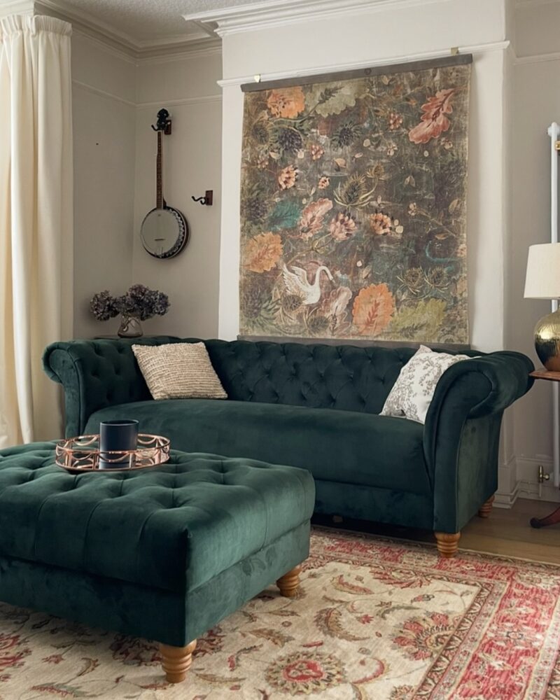 A sofa and footstool-living room furniture-green velvet chesterfield style sofa and ottoman footstool-vintage rug-ornate wall hanging-neutral walls-picture rail-cornicing-banjo-cream curtains-metal lamp-footstool styled as a coffee table with tray