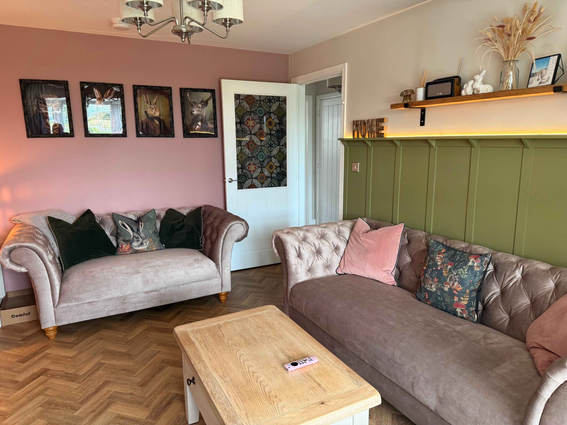 A couple of sofas and a coffee table-living room furniture-green panelled wall-pink feature wall-velvet mink coloured chesterfield style sofas-patterned cushions-velvet cushions-gallery wall-high wooden shelving-ceiling light-wooden and painted coffee table-parquet wooden floor