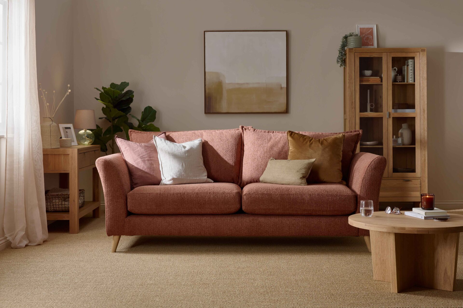 A sofa, coffee table, display cabinet, and console table-living room furniture-terracotta coloured sofa-wooden console table-neutral walls-sisal carpet-cream vase with dried flowers-potted house plant-framed photo-glass lamp-neutral abstract wall art-draped sheer curtain-wooden and glass display cabinet-round wooden coffee table