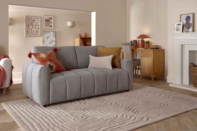 A sofa and desk-living room furniture-grey sofa bed-patterned cushion-neutral cushions-textured neutral rug-parquet wooden floor-wooden desk-fireplace-corridor-beige walls-framed abstract wall art-terracotta lamp-woven wood basket