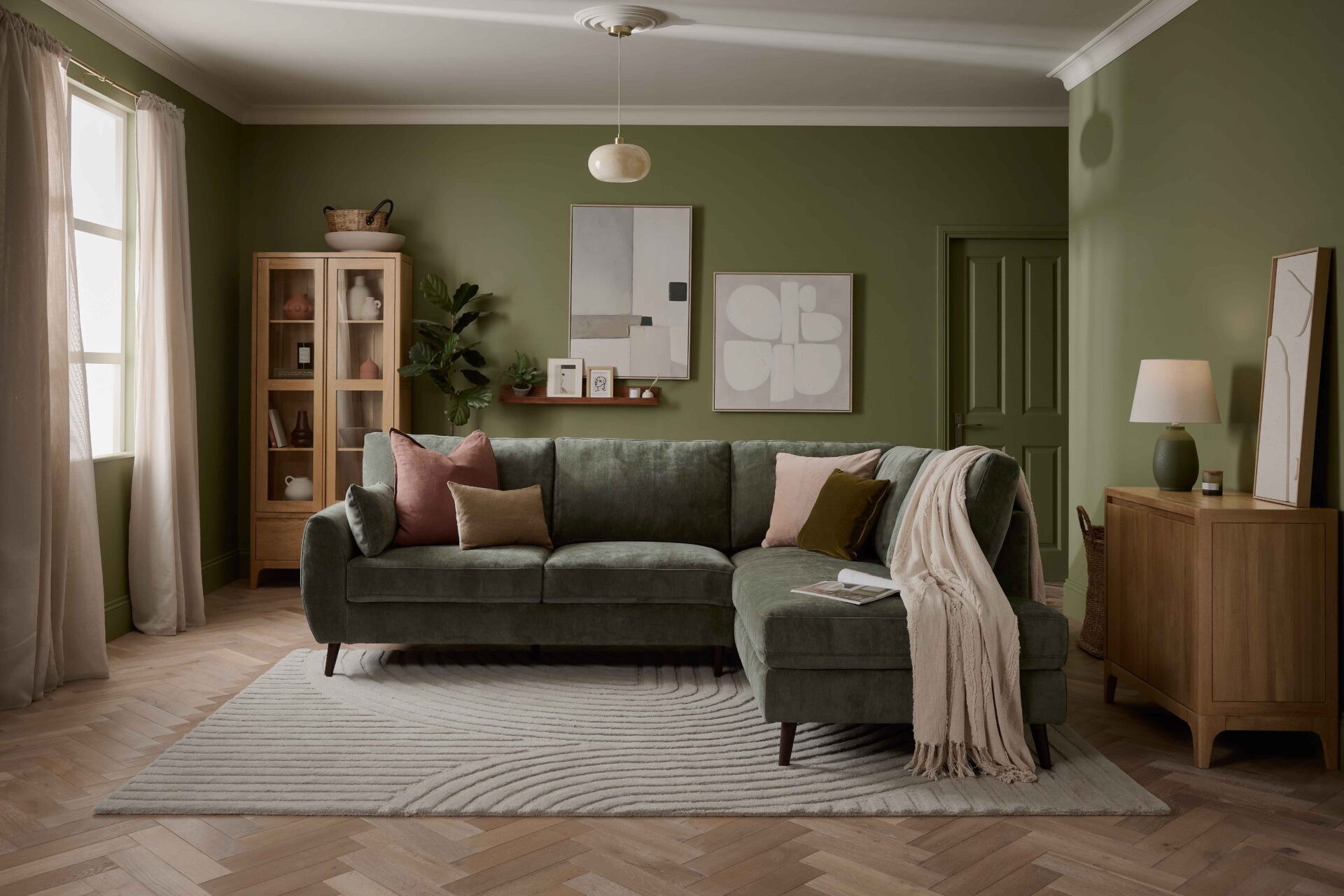 A sofa, sideboard, and display cabinet-living room furniture-green corner sofa-green walls-neutral ceiling-glass ceiling light-wooden sideboard-wood and glass display cabinet-neutral abstract wall art-floating wooden shelf-sheer draped curtains-textured neutral rug-wooden parquet floor-draped beige throw-scatter cushions