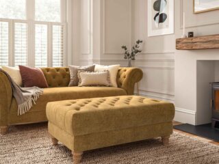 A sofa-living room furniture-yellow coloured chesterfield style sofa and ottoman footstool-neutral cushions-neutral walls with panelling and beading-abstract artwork-jute rug-fireplace-window shutters-radiator-neutral throw