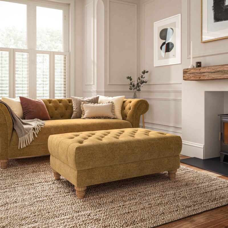 A sofa-living room furniture-yellow coloured chesterfield style sofa and ottoman footstool-neutral cushions-neutral walls with panelling and beading-abstract artwork-jute rug-fireplace-window shutters-radiator-neutral throw
