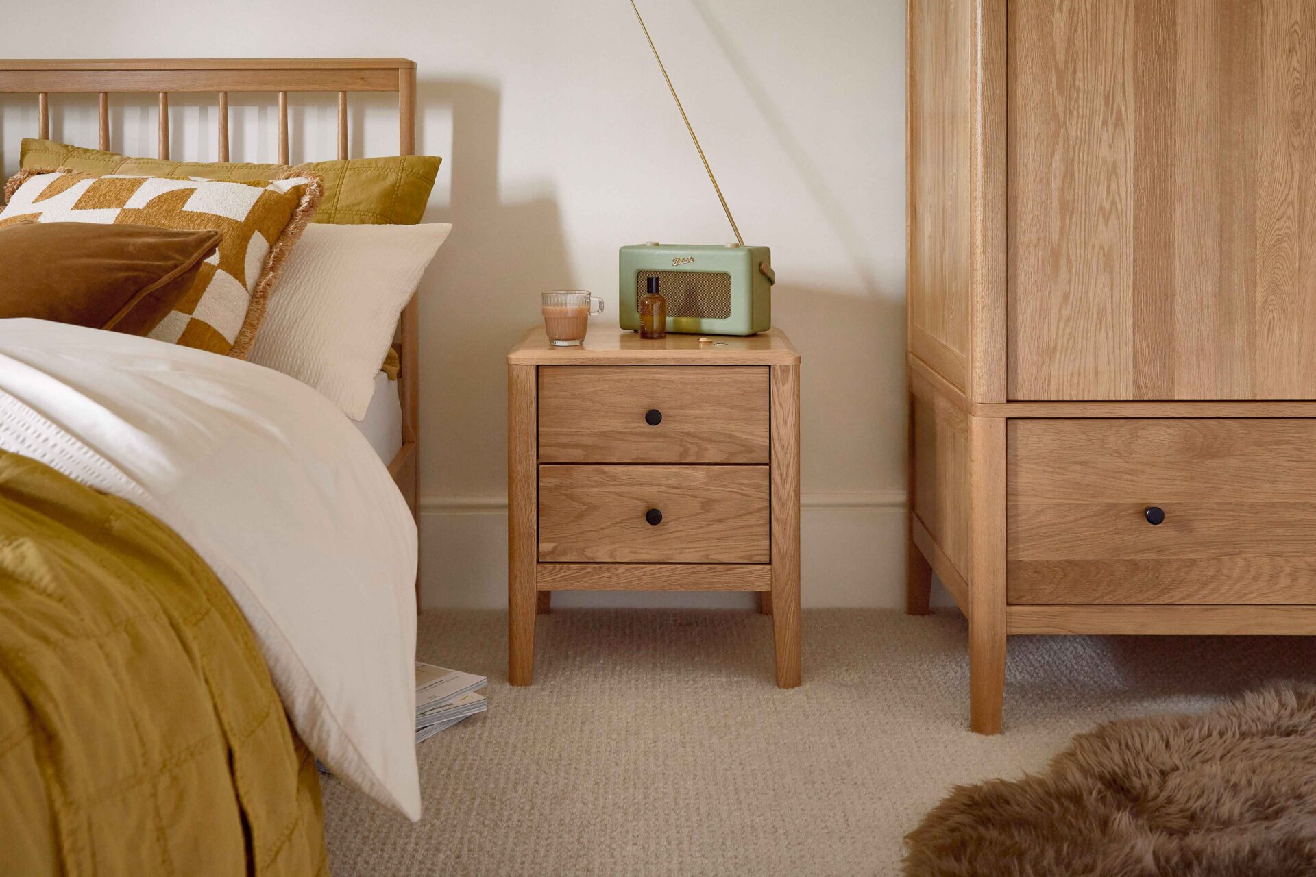 A bedside table, wardrobe, and bed-bedroom furniture-natural oak bedside table-natural oak wardrobe-wooden bed with spindle headboard-brown faux sheepskin rug-neutral carpet-cream walls-gold bedding-white bed linen-green retro radio-glass cup of tea
