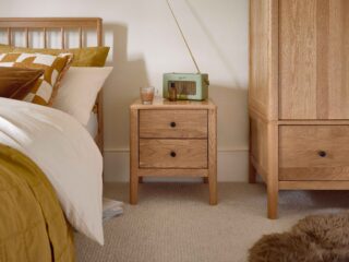 A bedside table, wardrobe, and bed-bedroom furniture-natural oak bedside table-natural oak wardrobe-wooden bed with spindle headboard-brown faux sheepskin rug-neutral carpet-cream walls-gold bedding-white bed linen-green retro radio-glass cup of tea