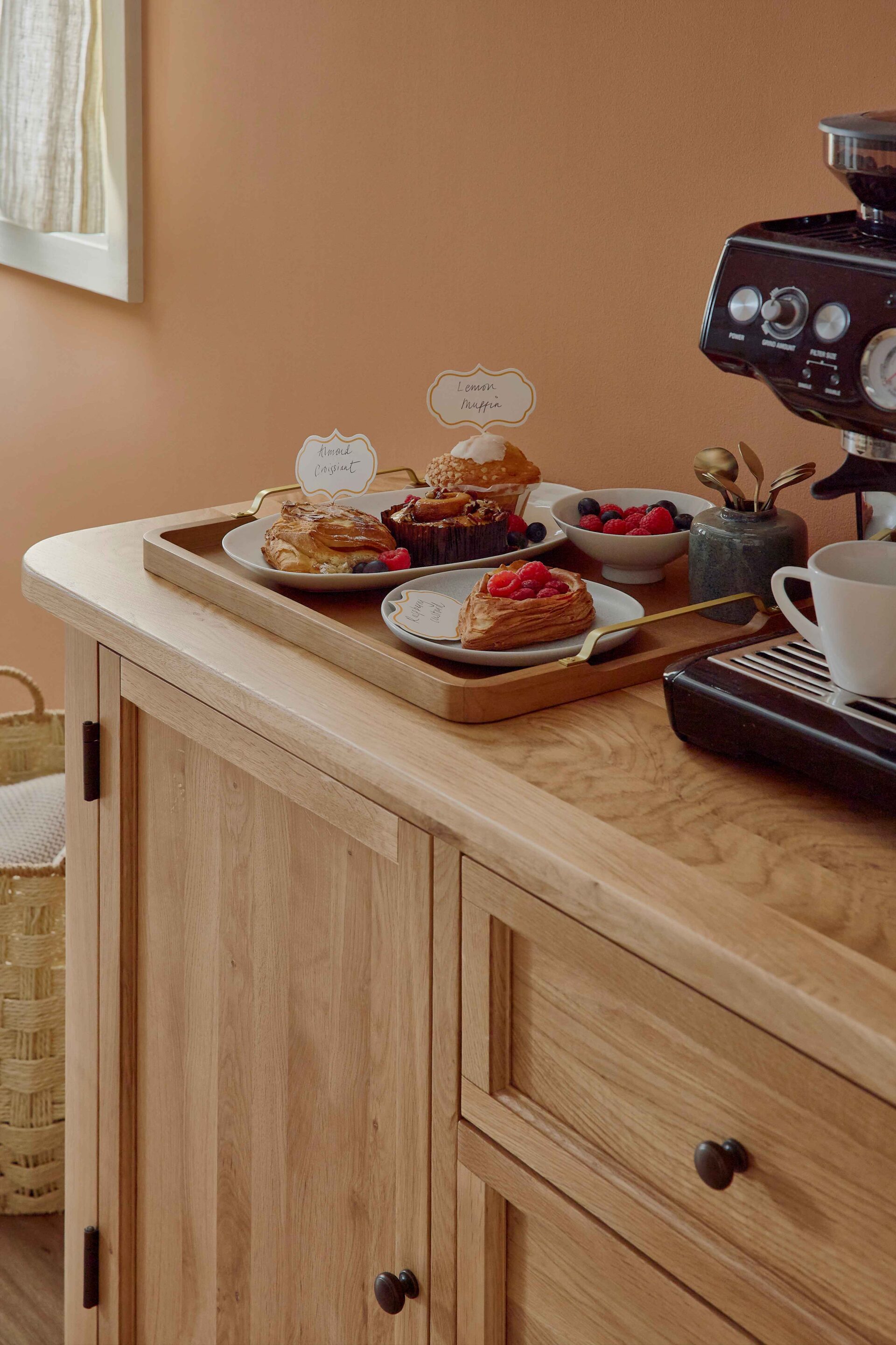 A sideboard-kitchen dining furniture-wooden sideboard-soft terracotta walls-black coffee machine-wooden tray-pastries-berries-serving platter-bowl-plate-pots of brass spoons-woven storage basket