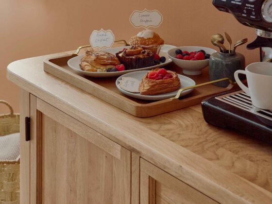 A sideboard-kitchen dining furniture-wooden sideboard-soft terracotta walls-black coffee machine-wooden tray-pastries-berries-serving platter-bowl-plate-pots of brass spoons-woven storage basket