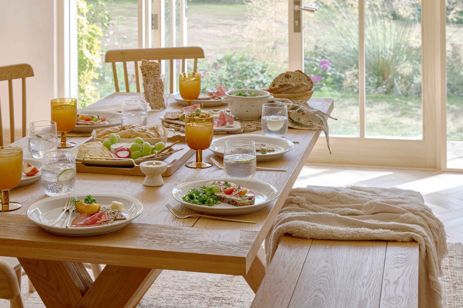 A dining table-dining room furniture-an oak dining table-oak bench-oak spindle back chairs-amber glassware-white tableware-gold cutlery-water glasses-food serving platter-salad-bread basket-wooden flooring-beige throw-jute rug-french doors-garden view