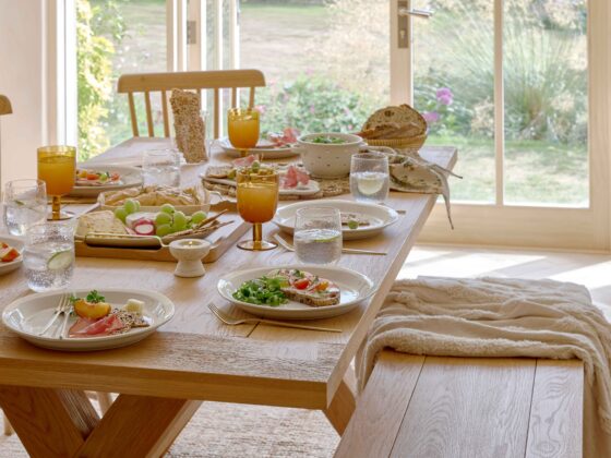 A dining table-dining room furniture-an oak dining table-oak bench-oak spindle back chairs-amber glassware-white tableware-gold cutlery-water glasses-food serving platter-salad-bread basket-wooden flooring-beige throw-jute rug-french doors-garden view