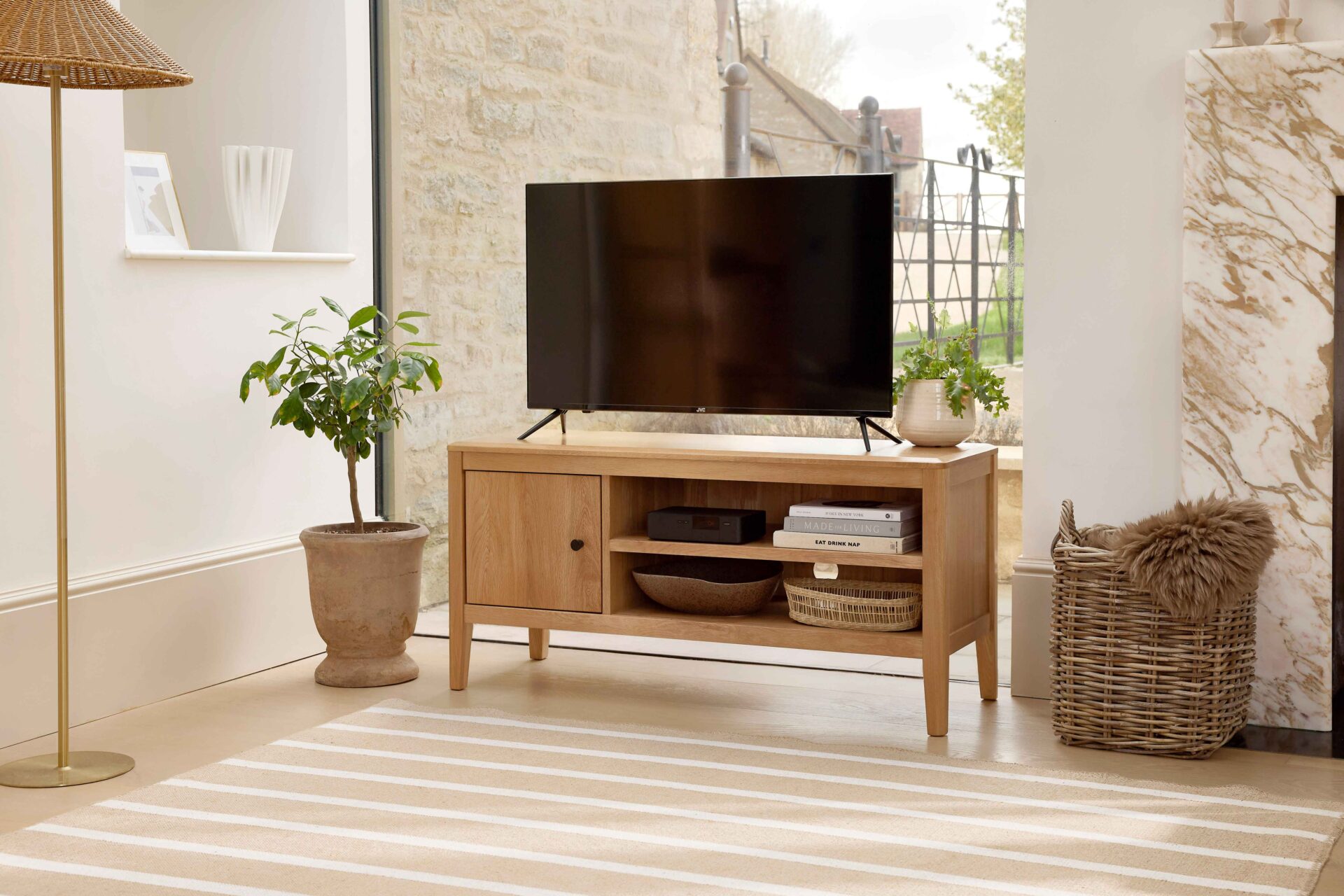 A TV unit-living room furniture-oak TV unit-striped jute rug-woven storage basket-faux brown sheepskin rug-clay pot with houseplant-floor standing rattan and brass lamp-marble fireplace-window wall-off white walls-white vase-stacked books-woven tray-TV