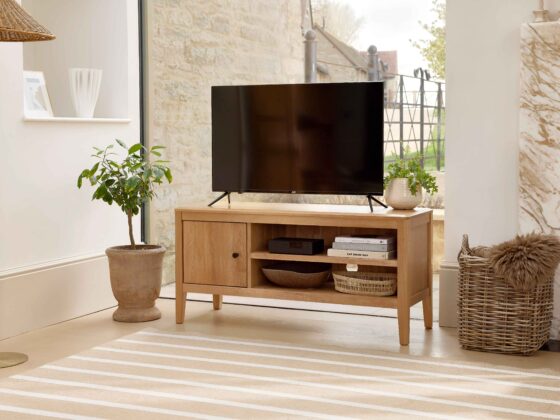 A TV unit-living room furniture-oak TV unit-striped jute rug-woven storage basket-faux brown sheepskin rug-clay pot with houseplant-floor standing rattan and brass lamp-marble fireplace-window wall-off white walls-white vase-stacked books-woven tray-TV