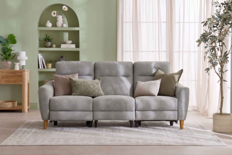 A sofa and console table-living room furniture-grey leather recliner sofa-earthy scatter cushions-vintage style rug-sheer curtains-sage green walls-built in arched shelving-oak console table-potted houseplant-cream vase with white flowers-woven basket-stacked books
