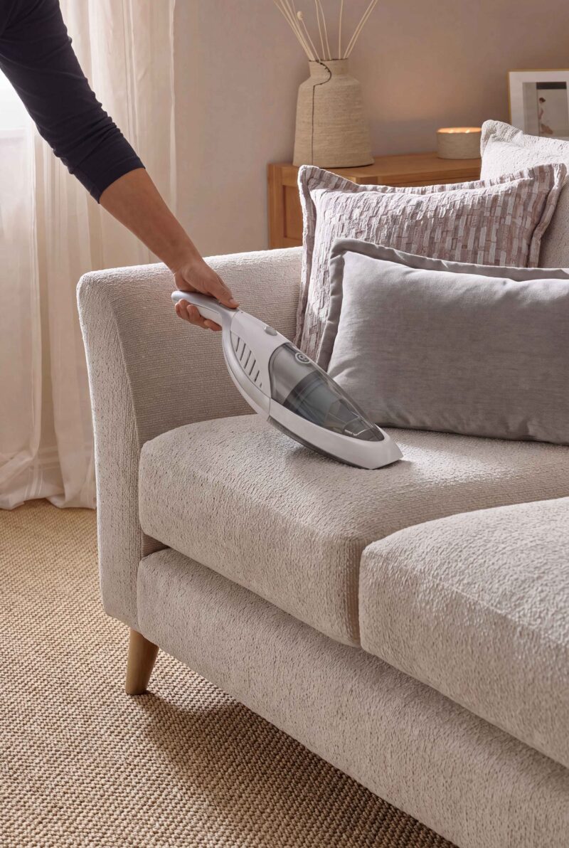 A sofa and vacuum cleaner-living room furniture-beige sofa-neural cushions-hand held small vacuum cleaner-sisal carpet-neutral walls-wooden sideboard-vase-sheer curtain-black top-arm