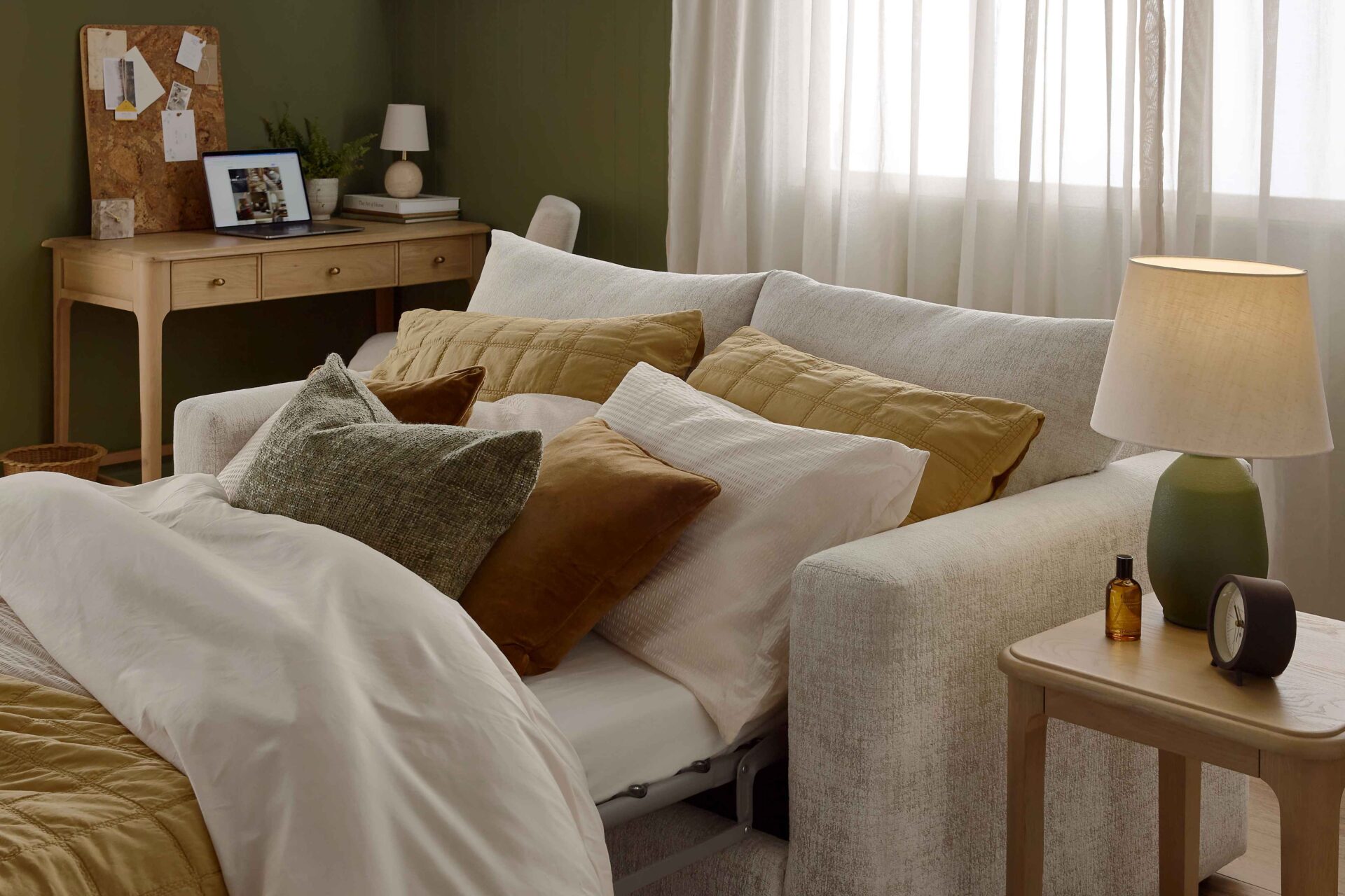 A sofa bed, side table, and desk-living room furniture-wooden side table-2 seater sofa bed-yellow bed linen-brown velvet cushions-green pillow-green table lamp-wooden desk with drawers-cork board-laptop-green walls-sheer curtains