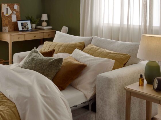 A sofa bed, side table, and desk-living room furniture-wooden side table-2 seater sofa bed-yellow bed linen-brown velvet cushions-green pillow-green table lamp-wooden desk with drawers-cork board-laptop-green walls-sheer curtains