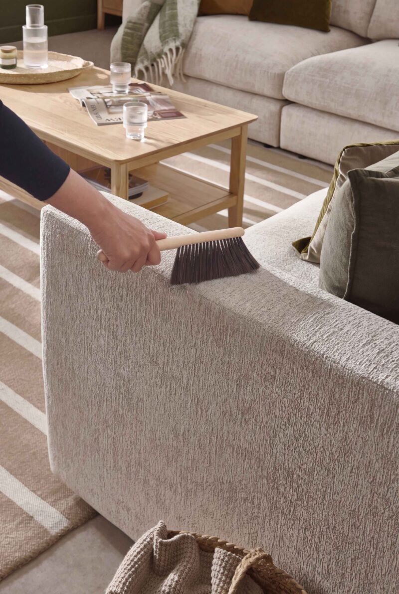 A sofa, coffee table, and brush-living room furniture-beige corner sofa-striped jute rug-wooden coffee table with shelf-black top-arm-wooden handled brush-water carafe and glasses-scatter cushions