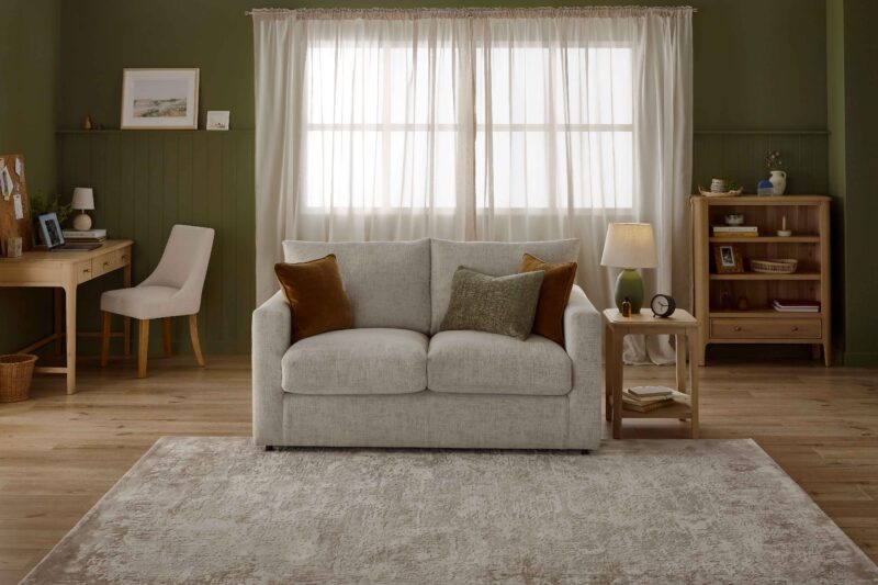 A sofa bed, side table, desk, and small bookcase-living room furniture-wooden side table-beige 2 seater sofa bed-brown velvet cushions-green cushion-green table lamp-wooden desk with drawers-cork board-laptop-green walls-sheer curtains-neutral textured rug- fabric desk chair-small wooden bookcase-stacked books