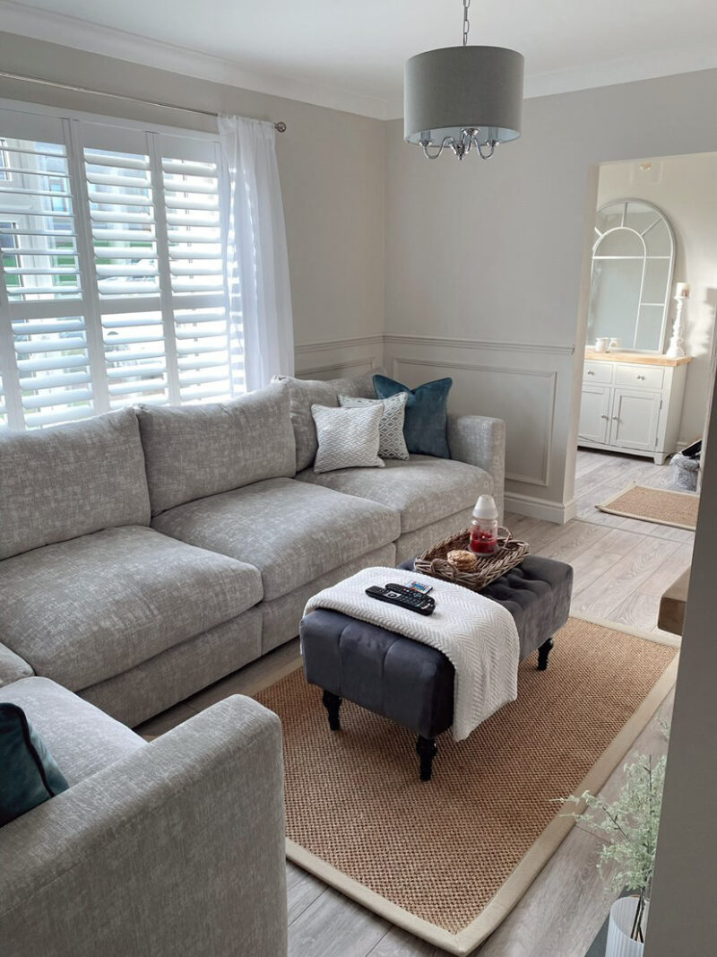 A corner sofa-living room furniture-large grey corner sofa-blue scatter cushions-sisal rug-pale wooden floor-neutral walls-charcoal footstool-knitted cream throw-white shutters-sheer curtains-woven tray-arched mirror-painted sideboard-grey ceiling light