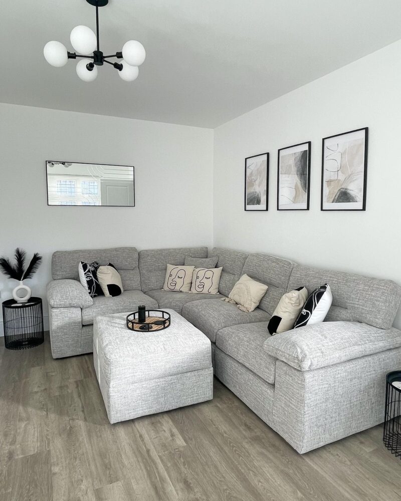 A corner sofa and footstool-living room furniture-pale grey modular corner sofa-pale grey chunky footstool-wooden flooring-black metal wire side tables-monochrome cushions-white walls-black framed abstract artwork-horizontal mirror-black and white ceiling light
