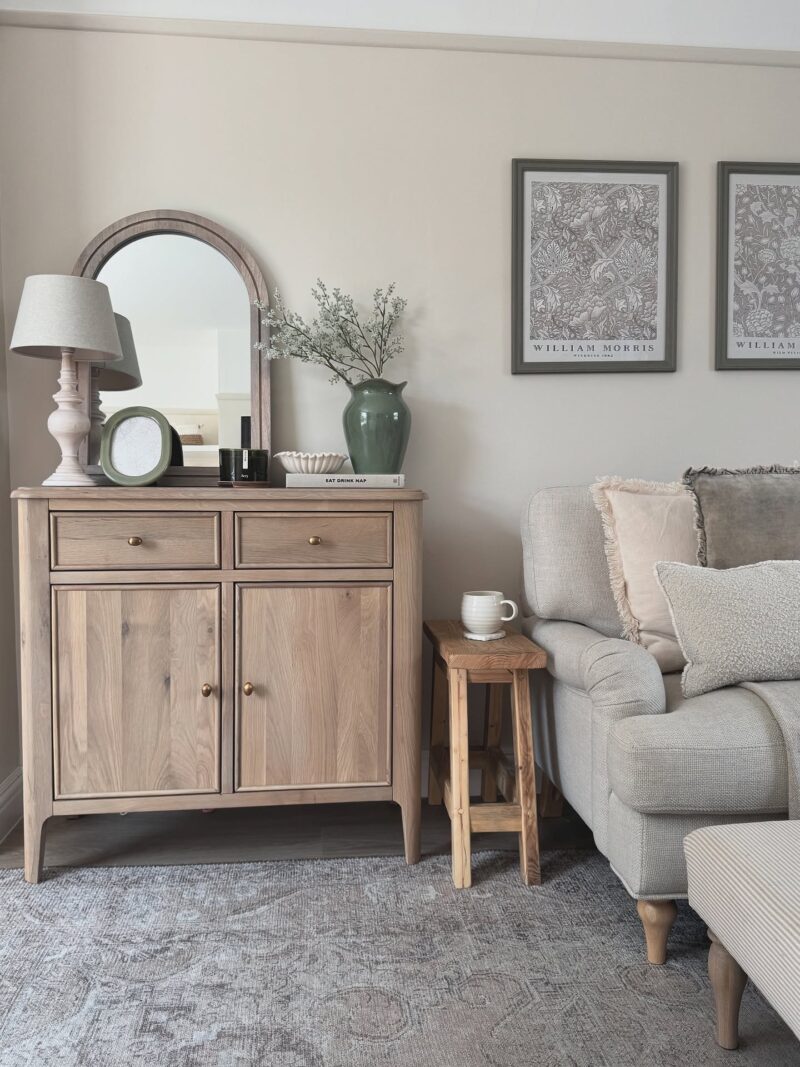 A sideboard and sofa-living room furniture-pale oak small sideboard-beige walls-grey rug-wooden side table-grey sofa-textured neutral scatter cushions-neutral striped footstool-framed william morris wall art-wooden arched mirror-neutral table lamp-green vase