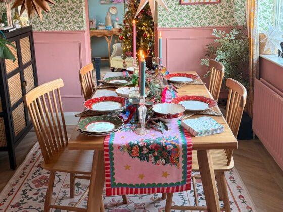 A dining table and chairs-dining room furniture-wooden dining table with play legs-spindle back wooden chairs-floral patterned rug-christmas table runner-pink and green tapered candles-christmas crockery-paper star decorations-pink painted panelling-green patterned wallpaper-black and rattan storage unit
